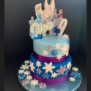 Op de afbeelding: Een tweelaagse blauwe en paarse taart versierd met sneeuwvlokkensprinkels en een Frozen-thema topper met Elsa, Anna en een kasteel. De taart is versierd met de naam "Harper" in gouden glitter.