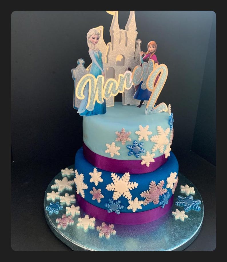 Op de afbeelding: Een tweelaagse blauwe en paarse taart versierd met sneeuwvlokkensprinkels en een Frozen-thema topper met Elsa, Anna en een kasteel. De taart is versierd met de naam "Harper" in gouden glitter.