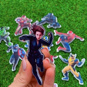 Puede incluir: Una colecci&oacute;n de pegatinas de personajes de superh&eacute;roes, incluyendo Viuda Negra, Iron Man, Spider-Man y Wolverine, se muestra sobre un fondo verde. La pegatina de Viuda Negra se sostiene en una mano, mostrando al personaje en una pose din&aacute;mica.