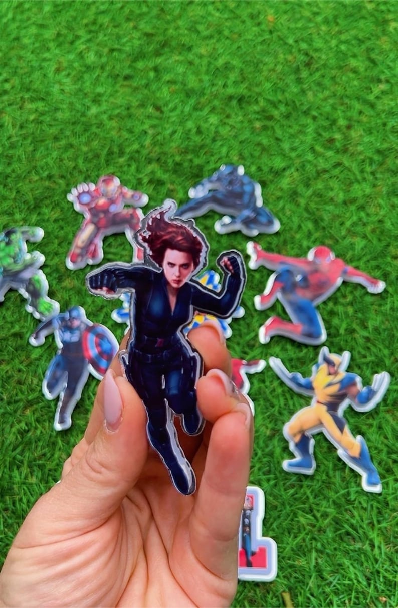 Puede incluir: Una colecci&oacute;n de pegatinas de personajes de superh&eacute;roes, incluyendo Viuda Negra, Iron Man, Spider-Man y Wolverine, se muestra sobre un fondo verde. La pegatina de Viuda Negra se sostiene en una mano, mostrando al personaje en una pose din&aacute;mica.