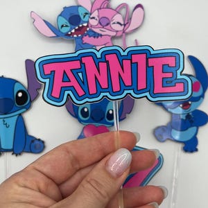 Puede incluir: Personajes de dibujos animados azules y rosas con el nombre "ANNIE" en letras rosas sobre un fondo azul. Los personajes sostienen un coraz&oacute;n y est&aacute;n en un adorno para pastel.
