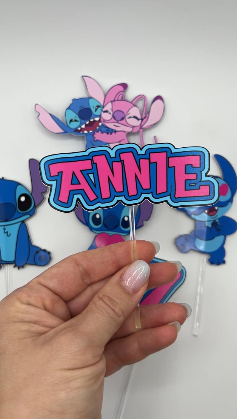 Puede incluir: Personajes de dibujos animados azules y rosas con el nombre "ANNIE" en letras rosas sobre un fondo azul. Los personajes sostienen un coraz&oacute;n y est&aacute;n en un adorno para pastel.
