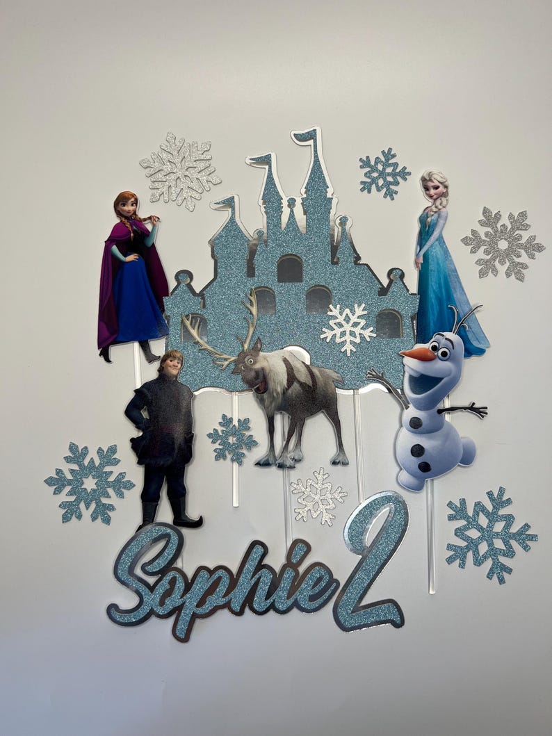 Op de afbeelding: Een blauwe en witte Frozen-taarttopper met een kasteel, Olaf, Elsa, Anna, Kristoff en Sven. De topper bevat ook sneeuwvlokken en de naam "Sophie 2".