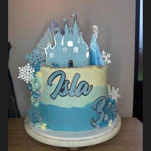 Op de afbeelding: Een blauwe en witte geglazuurde taart versierd met een glinsterend kasteel, een sneeuwvlokkenontwerp en een beeldje van Elsa uit Frozen. De taart is bedekt met een zilveren glitternaam "Isla" en het nummer "3" in zilveren glitter.