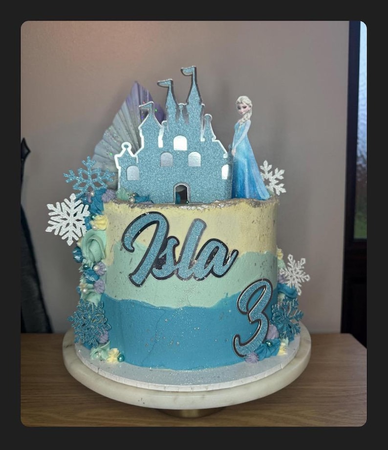 Op de afbeelding: Een blauwe en witte geglazuurde taart versierd met een glinsterend kasteel, een sneeuwvlokkenontwerp en een beeldje van Elsa uit Frozen. De taart is bedekt met een zilveren glitternaam "Isla" en het nummer "3" in zilveren glitter.