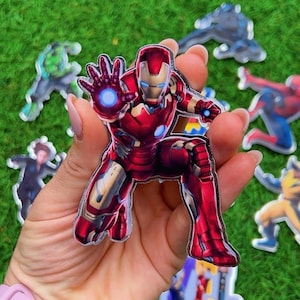 Puede incluir: Una pegatina de Iron Man roja y dorada en posici&oacute;n agachada, con una explosi&oacute;n de energ&iacute;a azul que emana de su mano. Otras pegatinas de superh&eacute;roes son visibles en el fondo sobre una superficie verde.