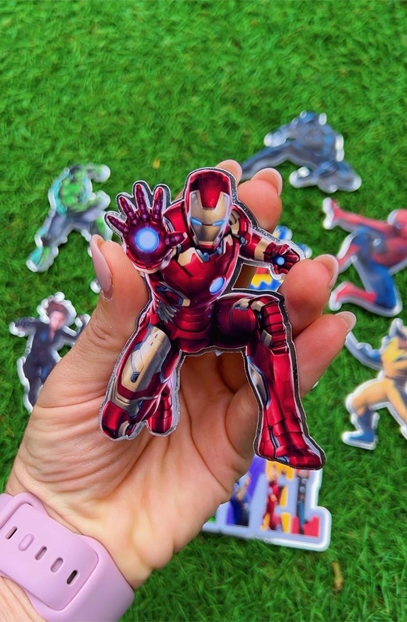 Puede incluir: Una pegatina de Iron Man roja y dorada en posici&oacute;n agachada, con una explosi&oacute;n de energ&iacute;a azul que emana de su mano. Otras pegatinas de superh&eacute;roes son visibles en el fondo sobre una superficie verde.