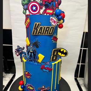 Puede incluir: Una tarta de cumplea&ntilde;os azul de dos pisos decorada con figuras de superh&eacute;roes, exclamaciones de c&oacute;mic y el nombre "Kairo". La tarta presenta adornos de Hulk, Capit&aacute;n Am&eacute;rica, Spider-Man y Batman. Gotas amarillas y el n&uacute;mero 8 completan el dise&ntilde;o.