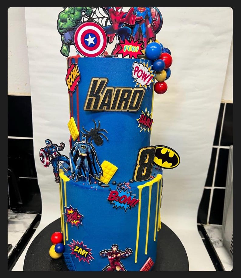 Puede incluir: Una tarta de cumplea&ntilde;os azul de dos pisos decorada con figuras de superh&eacute;roes, exclamaciones de c&oacute;mic y el nombre "Kairo". La tarta presenta adornos de Hulk, Capit&aacute;n Am&eacute;rica, Spider-Man y Batman. Gotas amarillas y el n&uacute;mero 8 completan el dise&ntilde;o.