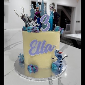 Op de afbeelding: Een gele cake met glazuur, versierd met een paars kasteel, een rendier, een sneeuwvlok, een paars cijfer 3, de naam "Ella" in paars en een paars en blauw Olaf-figuurtje.