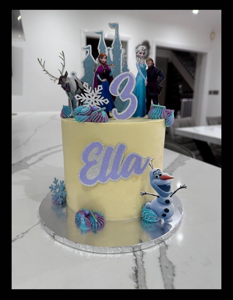 Op de afbeelding: Een gele cake met glazuur, versierd met een paars kasteel, een rendier, een sneeuwvlok, een paars cijfer 3, de naam "Ella" in paars en een paars en blauw Olaf-figuurtje.