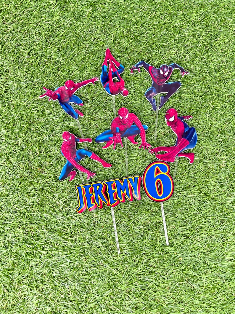 May include: A set of 7 cake toppers featuring Spiderman in various poses, a blue number 6, and the name "JEREMY" in red and yellow letters. The toppers are on sticks and are set against a green grass background.
