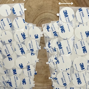 May include: A close-up of a pile of round, white, double-coated tissue tape circles with the 3M logo and the text "9448A Double Coated Tissue Tape" printed on them. The circles are arranged in a random pattern on a wooden surface. The tape is 23 mm in diameter and 28 mm in diameter.