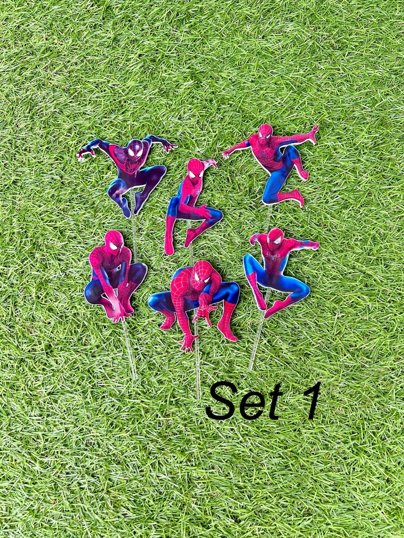 May include: Set of six red and blue Spiderman cake toppers. The toppers are on a green grass background. The text "Set 1" is visible in the bottom right corner of the image.