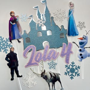 Op de afbeelding: Frozen-thema cake topper set met een blauw glitterkasteel, Elsa, Anna, Olaf, Sven en sneeuwvlokken. De set bevat ook een paarse glitter "Lola 4" naamtopper.