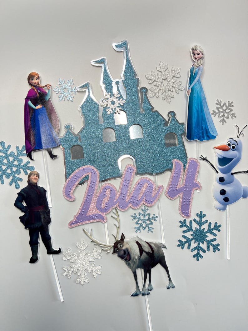 Op de afbeelding: Frozen-thema cake topper set met een blauw glitterkasteel, Elsa, Anna, Olaf, Sven en sneeuwvlokken. De set bevat ook een paarse glitter "Lola 4" naamtopper.