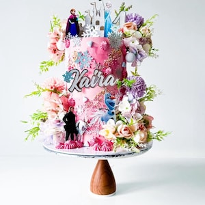 Op de afbeelding: Een roze geglazuurde taart versierd met bloemen, sneeuwvlokken en Frozen-figuren. De taart staat op een witte taartstandaard met een houten voet. De taart is versierd met de naam "Kaira".