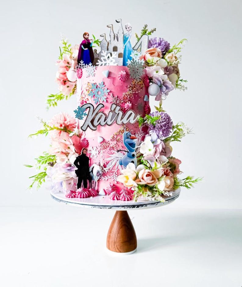 Op de afbeelding: Een roze geglazuurde taart versierd met bloemen, sneeuwvlokken en Frozen-figuren. De taart staat op een witte taartstandaard met een houten voet. De taart is versierd met de naam "Kaira".