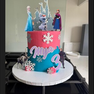 Op de afbeelding: Een tweelaagse verjaardagstaart versierd met een Frozen-thema. De taart is geglazuurd in roze en blauw met witte sneeuwvlokkensprinkels. De bovenste laag heeft een kasteel, Olaf, Elsa en Anna. De onderste laag heeft de naam "Mya" en het nummer "3" in paarse glitter.