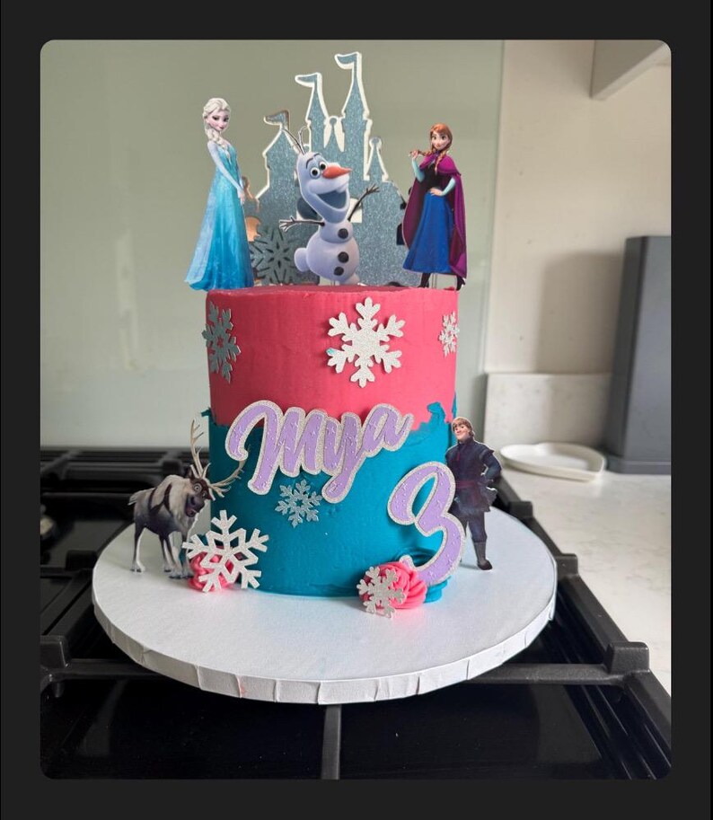 Op de afbeelding: Een tweelaagse verjaardagstaart versierd met een Frozen-thema. De taart is geglazuurd in roze en blauw met witte sneeuwvlokkensprinkels. De bovenste laag heeft een kasteel, Olaf, Elsa en Anna. De onderste laag heeft de naam "Mya" en het nummer "3" in paarse glitter.