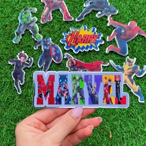 Puede incluir: Una colecci&oacute;n de pegatinas de personajes de Marvel, incluyendo Iron Man, Spider-Man y Capit&aacute;n Am&eacute;rica, dispuestas sobre una superficie verde. La palabra "MARVEL" est&aacute; escrita con im&aacute;genes de personajes. Tambi&eacute;n hay una pegatina con el nombre "HARRY".