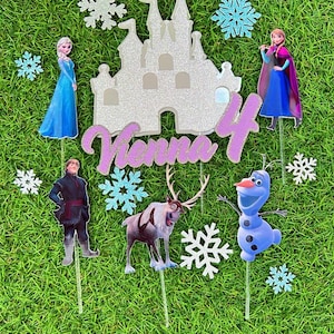 Op de afbeelding: Frozen-thema taarttoppers met Elsa, Anna, Olaf, Sven en een kasteel. De toppers zijn op stokjes en hebben een glinsterende afwerking. De naam "Venna 4" is in roze glitter op het kasteel geschreven.