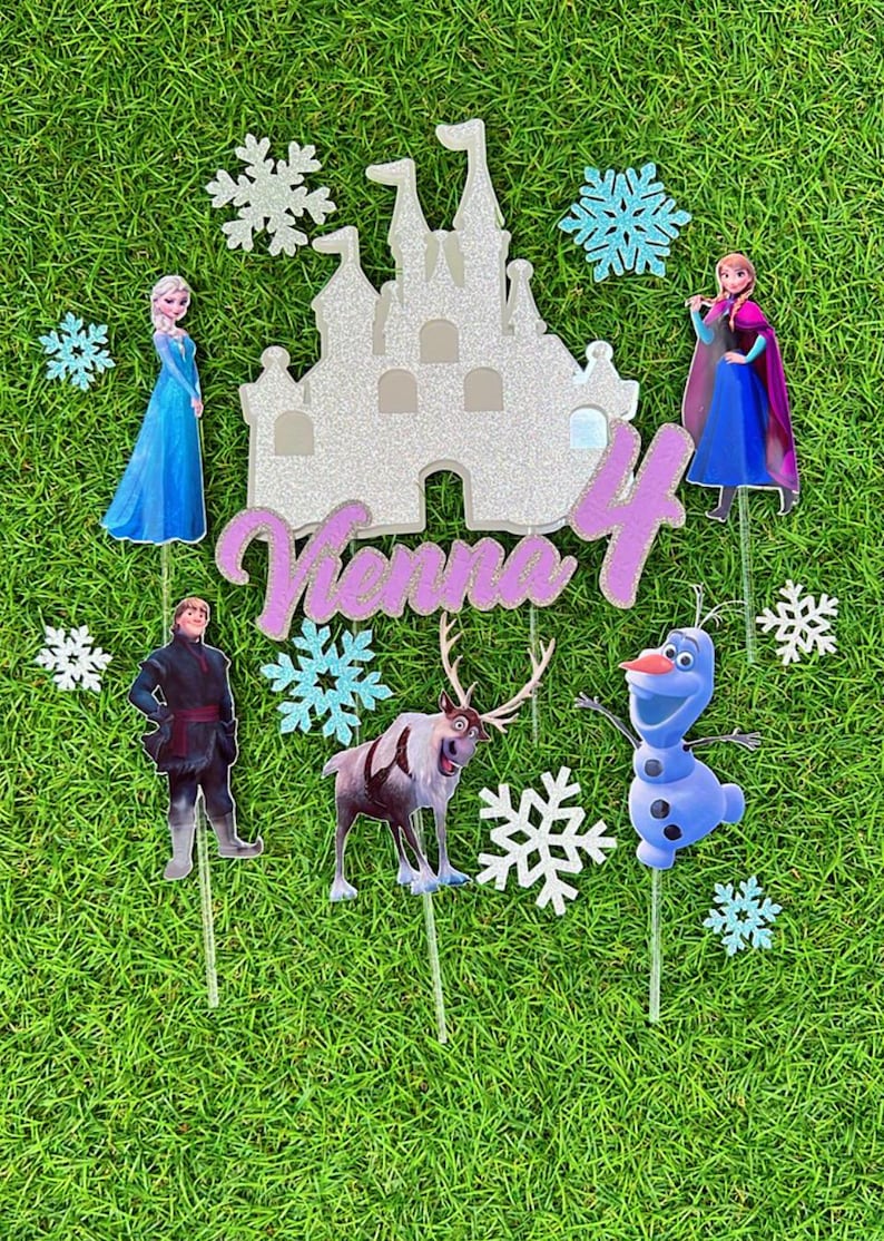 Op de afbeelding: Frozen-thema taarttoppers met Elsa, Anna, Olaf, Sven en een kasteel. De toppers zijn op stokjes en hebben een glinsterende afwerking. De naam "Venna 4" is in roze glitter op het kasteel geschreven.