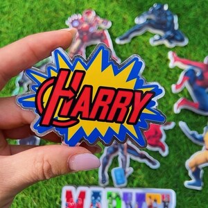 Puede incluir: Una pegatina transparente con el nombre "Harry" en rojo, dentro de un dise&ntilde;o de explosi&oacute;n amarillo y azul. Se ven otras pegatinas de personajes de Marvel y el logotipo de "Marvel" sobre un fondo verde, con tem&aacute;tica de superh&eacute;roes.