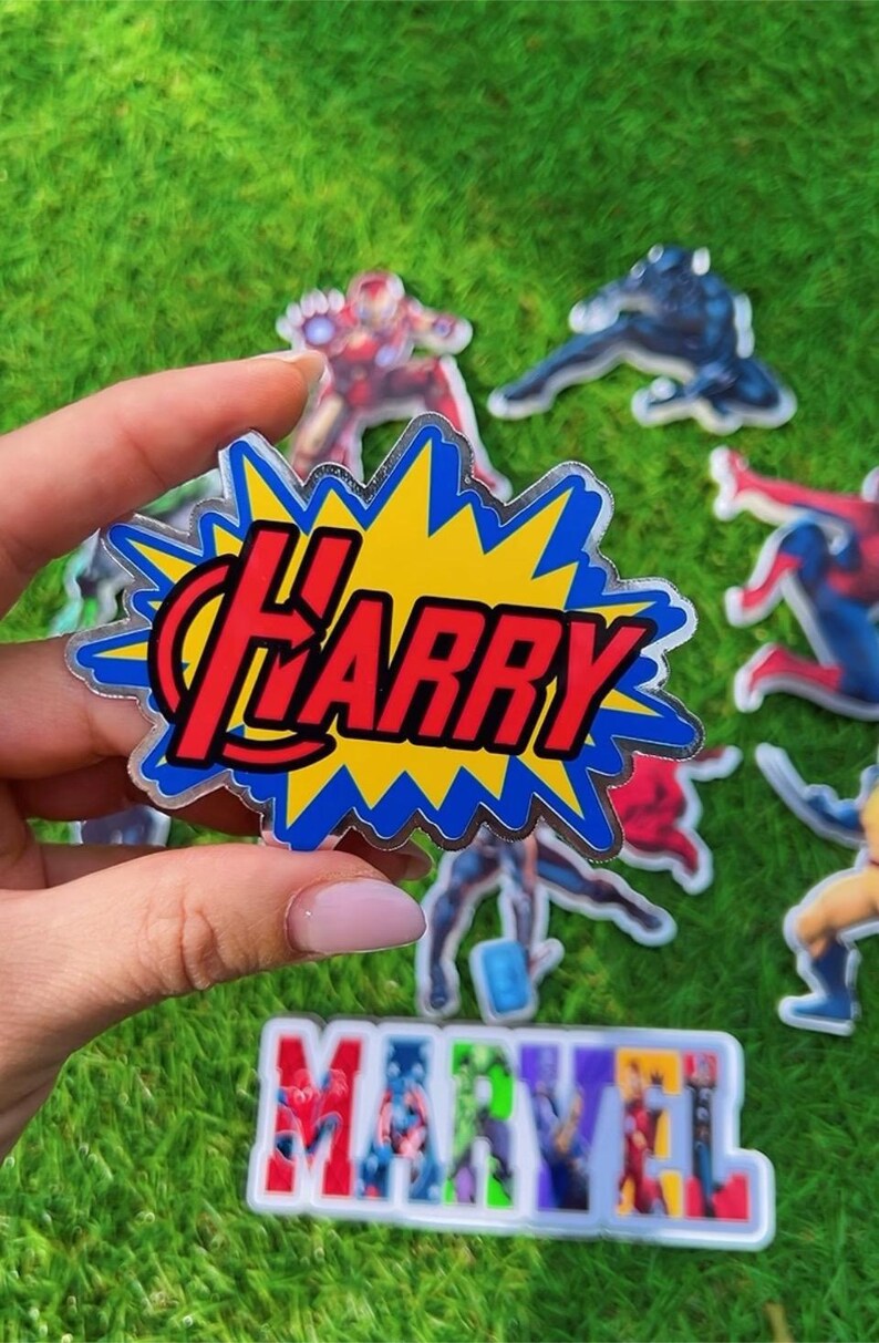 Puede incluir: Una pegatina transparente con el nombre "Harry" en rojo, dentro de un dise&ntilde;o de explosi&oacute;n amarillo y azul. Se ven otras pegatinas de personajes de Marvel y el logotipo de "Marvel" sobre un fondo verde, con tem&aacute;tica de superh&eacute;roes.