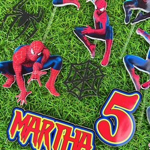 May include: A set of red and blue Spiderman cake toppers with black web designs. The toppers include a Spiderman character, a web, and the name "MARTHA" and the number "5".