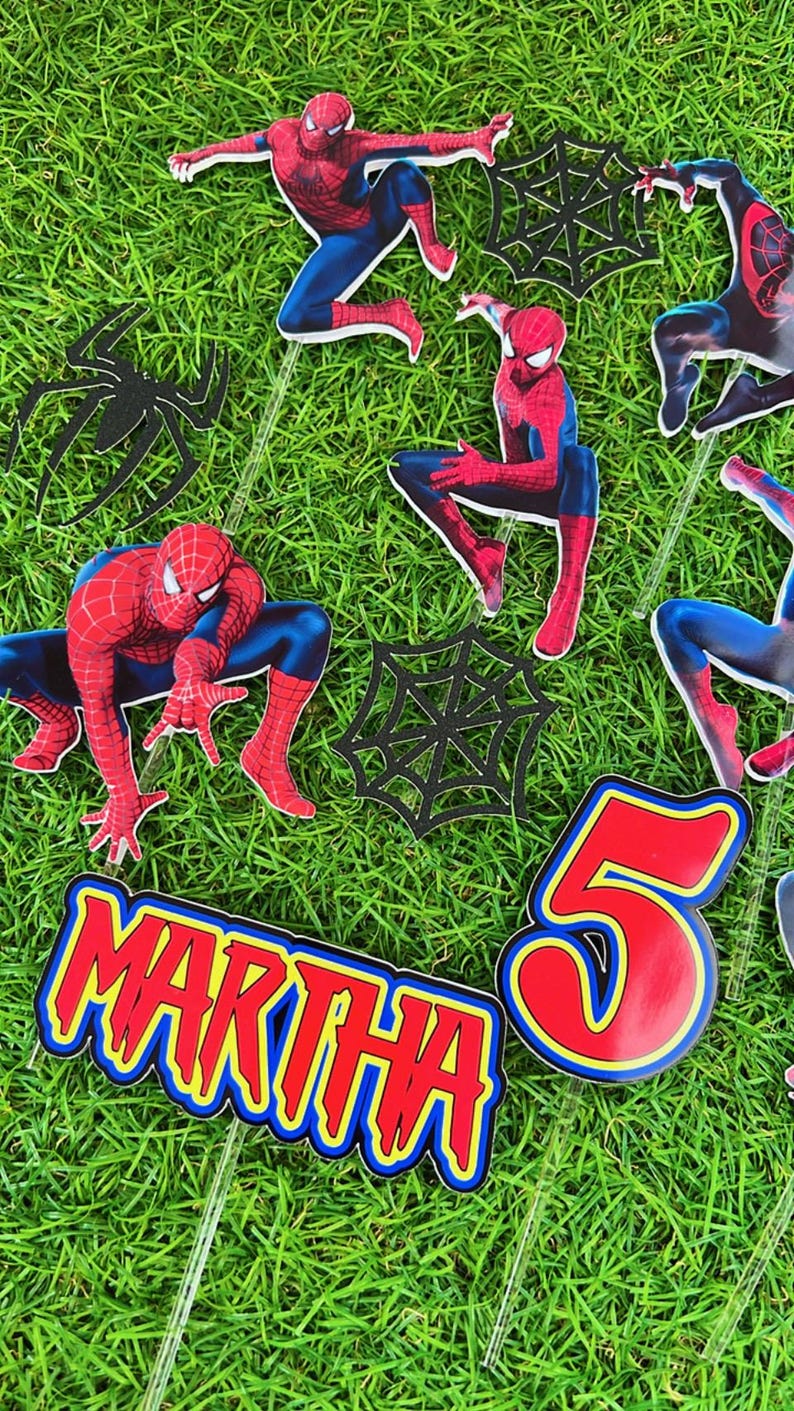 May include: A set of red and blue Spiderman cake toppers with black web designs. The toppers include a Spiderman character, a web, and the name "MARTHA" and the number "5".