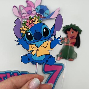 Puede incluir: Un personaje azul Stitch con una corona de flores y una camisa amarilla, levantando los brazos. El personaje est&aacute; en un adorno de pastel rosa y azul con el n&uacute;mero 7.