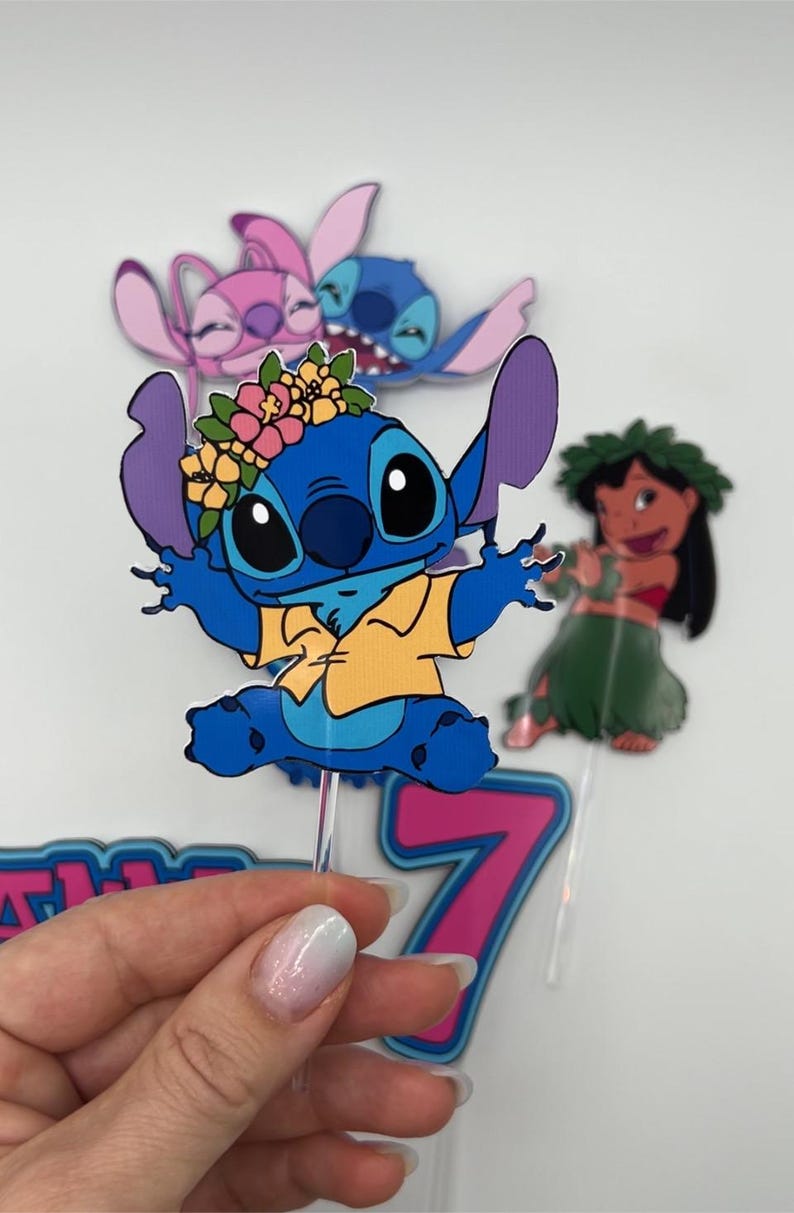 Puede incluir: Un personaje azul Stitch con una corona de flores y una camisa amarilla, levantando los brazos. El personaje est&aacute; en un adorno de pastel rosa y azul con el n&uacute;mero 7.