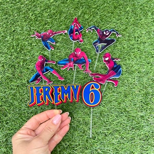 May include: A set of six cake toppers featuring Spiderman in various poses. The toppers are printed on cardstock and have a glossy finish. The set includes a topper with the name "JEREMY" and a topper with the number "6".