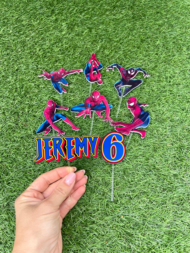 May include: A set of six cake toppers featuring Spiderman in various poses. The toppers are printed on cardstock and have a glossy finish. The set includes a topper with the name "JEREMY" and a topper with the number "6".