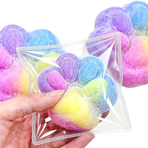 May include: A soft, plush paw-shaped toy featuring a vibrant tie-dye pattern of pink, yellow, blue, and purple. The toy is encased in a clear, square plastic bag, with additional identical paw-shaped toys visible in the background.