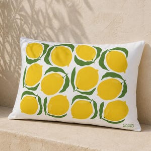 May include: White rectangular pillow with a repeating pattern of yellow lemons and green leaves. The pillow is on a stone surface next to a potted plant. The text "MAISON BOHO" is printed on the bottom right corner of the pillow.