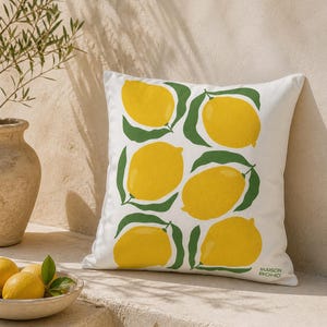 May include: White square throw pillow with a repeating pattern of yellow lemons and green leaves. The pillow has the text "MAISON BOHO" in green on the lower right corner. The pillow is on a stone surface.