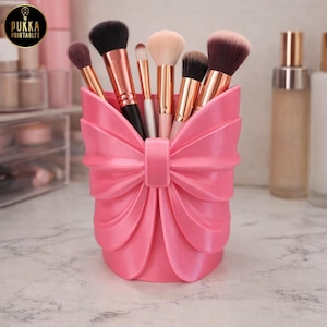 May include: A pink bow-shaped makeup brush holder filled with various makeup brushes. The holder is made of a solid pink material and has a bow design. The brushes have black, brown, and beige bristles with rose gold handles. The logo "PUKKA PRINTABLES" is in the top left corner.