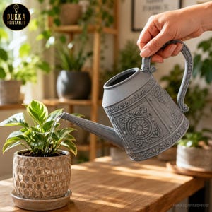 Puede incluir: Una regadera de plástico gris mate con un patrón floral y circular en relieve en el exterior. La regadera cuenta con un asa curva y un pico largo y delgado, vertiendo agua sobre una pequeña planta verde colocada en una maceta texturizada.