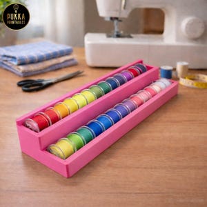 Puede incluir: Un organizador de hilos de madera rosa lleno de coloridas bobinas de hilo. Las bobinas están dispuestas en un patrón de arcoíris. Una máquina de coser y otros suministros de costura están en el fondo.
