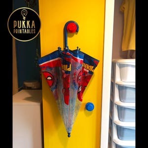 May include: A Spider-Man themed umbrella with a red, blue, and white design, hanging on a yellow wall. The umbrella is partially open, showing the Spider-Man logo and comic book graphics. A blue hook holds the umbrella.