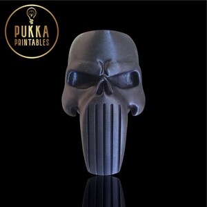 Op de afbeelding: Een zwart, schedelvormig decoratief item met een verticaal, geribbeld ontwerp onder de schedel. Het item heeft een metallic glans en staat tegen een zwarte achtergrond. Het logo "PUKKA PRINTABLES" staat linksboven.