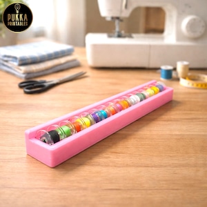 May include: A pink bobbin holder containing a variety of thread spools in clear casings. Sewing equipment, including scissors, fabric, and a sewing machine, are visible in the background. The logo "Pukka Printables" is in the upper left corner.