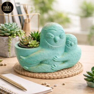 May include: A turquoise ceramic planter shaped like a mother sloth embracing a baby sloth. The planter holds several small succulents. The planter sits on a round woven mat. The logo "Pukka Printables" is visible.