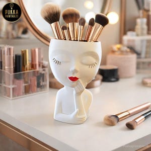 May include: A white ceramic makeup brush holder shaped like a woman's face with closed eyes and red lipstick. The holder is filled with various makeup brushes. The Pukka Printables logo is in the top left corner and the bottom right corner.