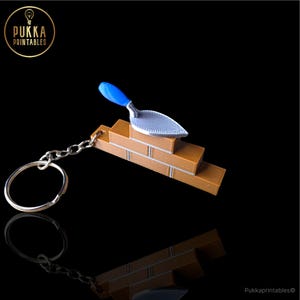 May include: A construction-themed keychain featuring a miniature brick wall and a trowel. The brick wall is brown with white mortar lines, and the trowel has a blue handle and a silver blade. The keychain includes a silver chain and ring.