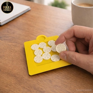 May include: A yellow tray holds eleven cream-colored, circular game pieces with star and snowflake patterns. One piece is being held by a hand. The tray and pieces are on a wooden surface. The text "Pukka Printables" is visible.