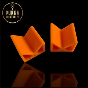 May include: Two bright orange, V-shaped bookends. The bookends are made of a solid material and have a geometric design. The logo "PUKKA PRINTABLES" is in a gold circle in the upper left corner.