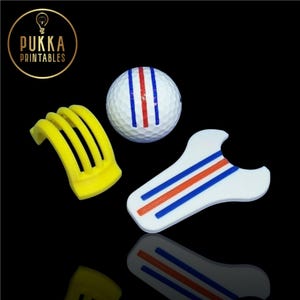 Peut inclure: Trois accessoires de golf imprimés en 3D : une balle de golf blanche avec des rayures rouges et bleues, un tee de golf jaune et un outil d’alignement de golf blanc avec des rayures rouges et bleues.